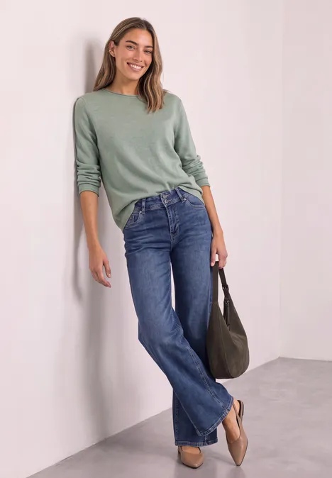 Jeune femme souriante appuyée sur un mur blanc, pull vert, jean bleu et sac à main.