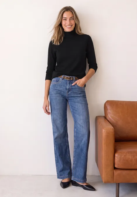 Eenvoudige trui met col Black Een vrouw in een zwarte coltrui en blauwe jeans staat nonchalant naast een bruine leren bank.