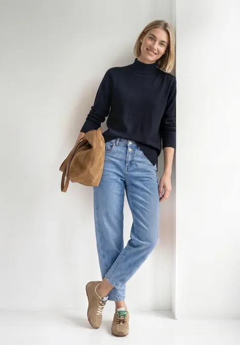 Basic Pullover mit Turtle Neck darkest blue Frau lehnt lächelnd an weißer Wand, trägt schwarzen Rollkragenpullover, blaue Jeans, hält beige Wildlederjacke.