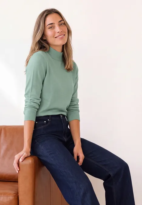 Basic Pullover mit Turtle Neck eucalyptus green Frau in grünem Rollkragenpullover und dunklen Jeans sitzt lächelnd auf einem braunen Sofa.