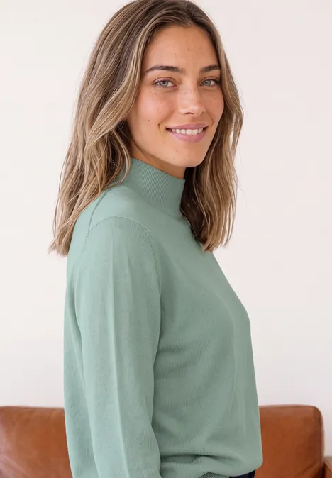 Basic Pullover mit Turtle Neck eucalyptus green Junge Frau lächelt, trägt grünen Rollkragenpullover, vor hellem Hintergrund.