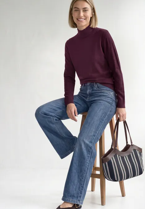 Basic Pullover mit Turtle Neck berry maroon Frau mit kurzem Blondhaar in lila Rollkragenpullover und ausgestellten Jeans sitzt auf Hocker, hält gestreifte Tasche