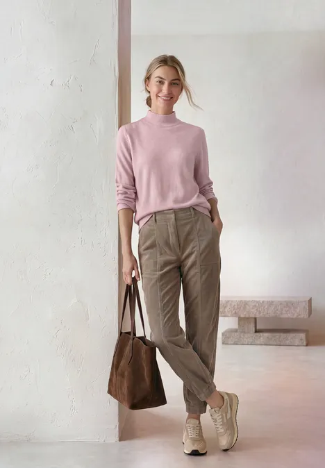 Basic Pullover mit Turtle Neck quiet rose mel. Frau in pastellrosa Pullover, beiger Hose, leichten Stiefeln und brauner Tasche vor schlichtem Hintergrund.