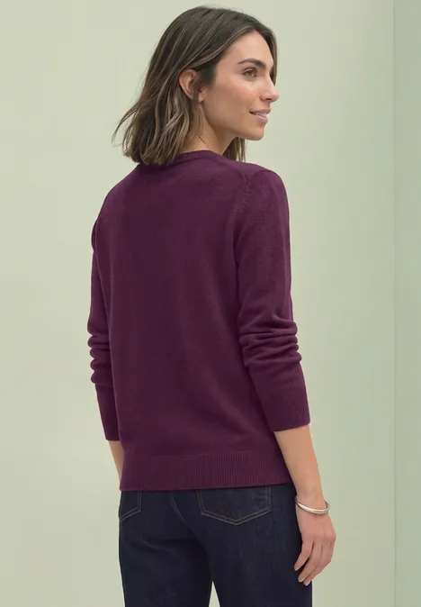 Frau in pflaumenfarbenem Pullover und dunkler Jeans, Rückenansicht vor neutraler Wand.