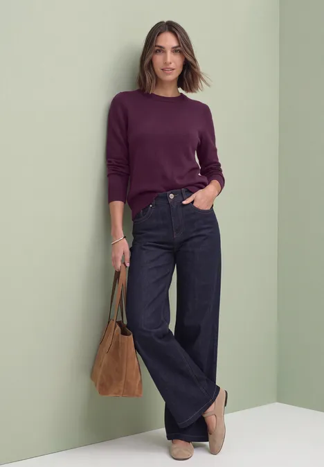 Frau in burgunderrotem Pullover, weiten Jeans, beigen Loafers und mit brauner Tasche vor hellem Hintergrund.
