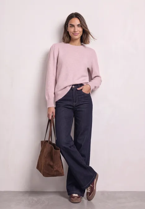 Frau in rosa Pullover, weiten Jeans und braunen Loafers trägt eine braune Tasche, posiert vor neutralem Hintergrund.