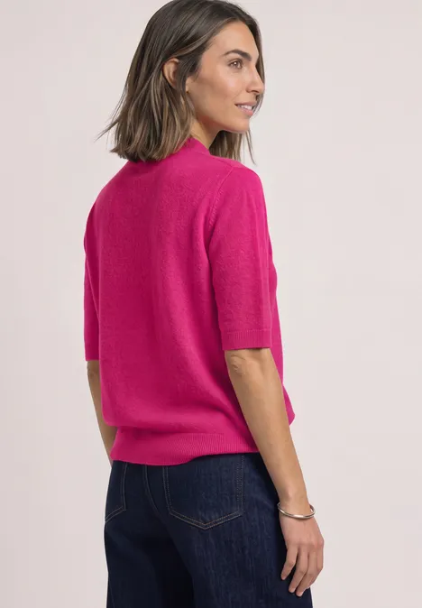 Frau mit schulterlangen Haaren trägt pinkfarbenen Kurzarm-Pullover und blaue Jeans, blickt seitlich.