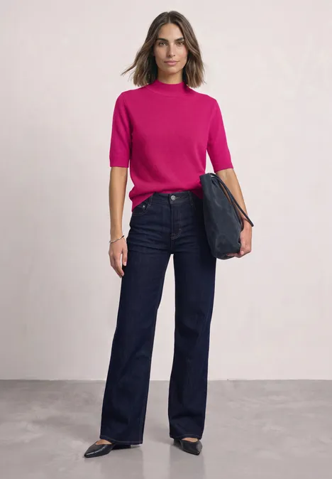 Eine Frau trägt ein pinkes Oberteil, weite Jeans und schwarze Schuhe, sie hält eine Clutch.