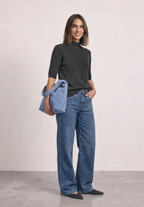 Frau in weiten Jeans und schwarzem Pulli, hellblaue Jacke lässig über der Schulter, lächelnd.