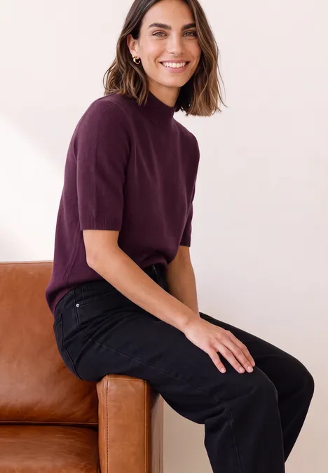 Eine lächelnde Frau sitzt auf der Sofalehne, trägt ein kurzärmeliges violettes Oberteil und schwarze Hose.