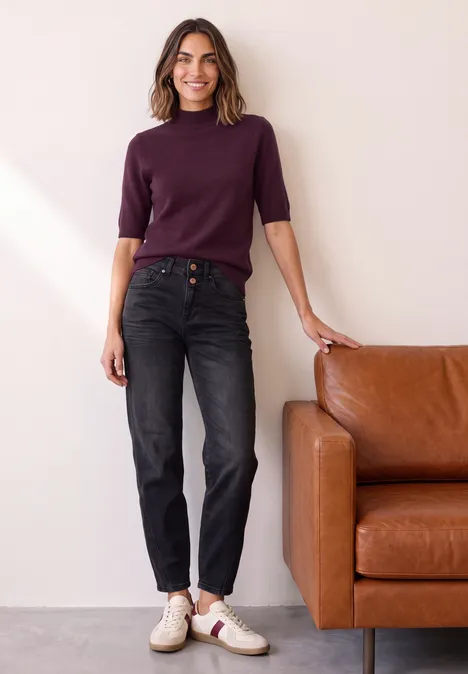 Frau lehnt an braunem Ledersofa, trägt lila Pullover, schwarze Jeans und weiße Sneaker und lächelt.