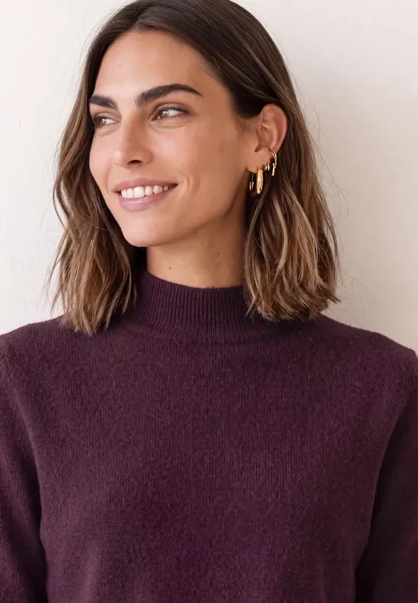 Frau mit dunkelbraunen Haaren und goldenen Ohrringen, lächelt in einem violetten Rollkragenpullover.