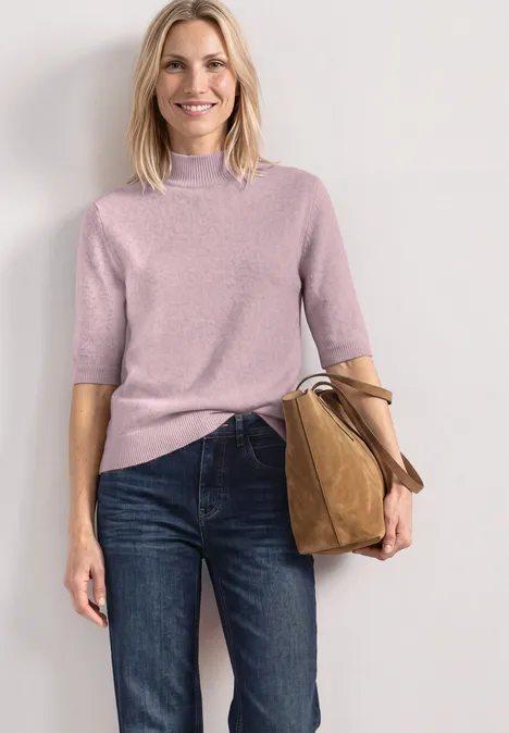 Eine Frau in rosa Kurzarmpullover, Jeans und trägt eine braune Tasche, steht vor neutralem Hintergrund.