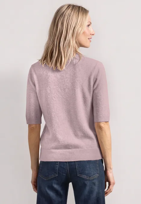 Frau mit schulterlangem blondem Haar trägt rosafarbenen Pullover und Blue Jeans, von hinten fotografiert.