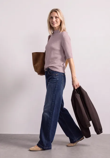 Frau in hellem Pullover und weitem Jeans, mit Tasche über der Schulter und Jacke am Arm.