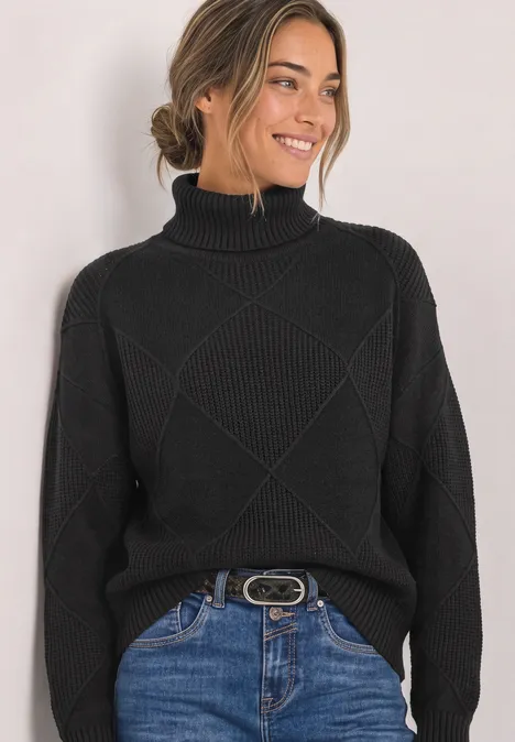 Frau trägt schwarzen Rollkragenpullover, Jeans mit Gürtel, lächelnd vor hellem Hintergrund.