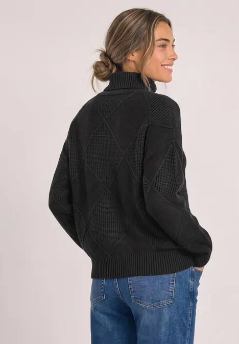 Frau in schwarzem Zopfmuster-Pullover und Jeans, seitlich stehend, lächelt, Haare lässig zum Dutt.