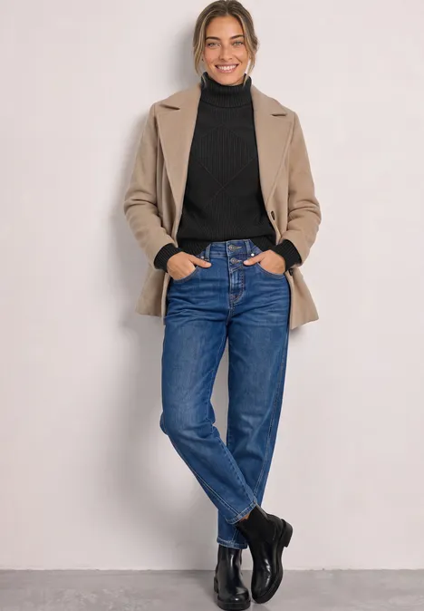 Frau in beigem Blazer, schwarzem Rollkragenpullover, blauen Jeans und schwarzen Schuhen posiert entspannt.