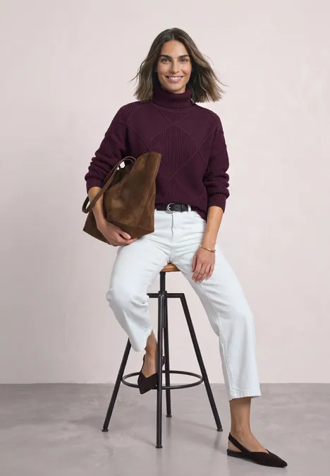 Eine Frau sitzt auf einem Hocker, trägt einen burgunderfarbenen Pullover und eine weiße Hose und hält eine braune Tasche.
