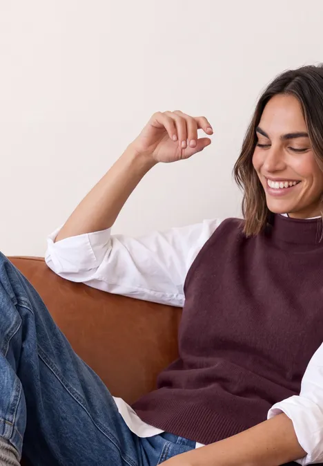 Eine lächelnde Frau entspannt sich auf einem Sofa, trägt Weste und Jeans.