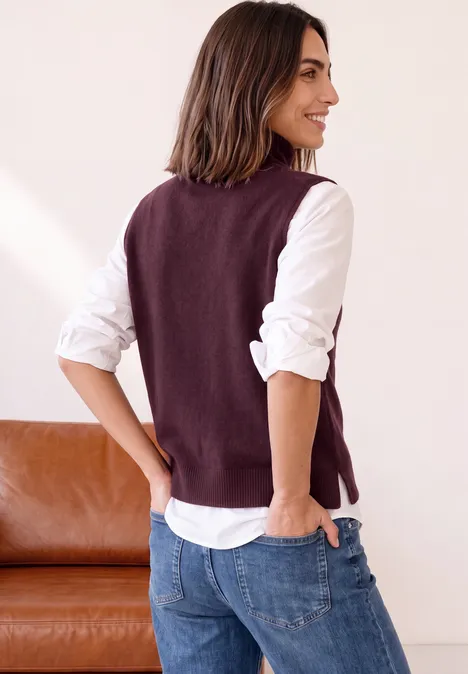 Frau trägt weiße Bluse, violette Strickweste und Jeans, schaut lächelnd seitlich.
