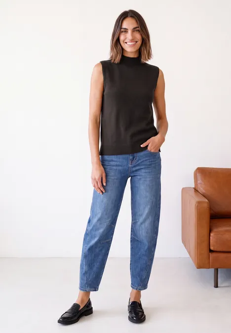 Eine lächelnde Frau in schwarzem ärmellosem Top, blauen Jeans und schwarzen Loafers steht vor einem braunen Ledersofa.