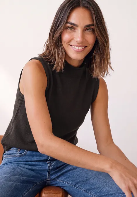 Eine lächelnde Frau mit dunkelbraunen Haaren, schwarzem ärmellosen Top und Jeans sitzt.