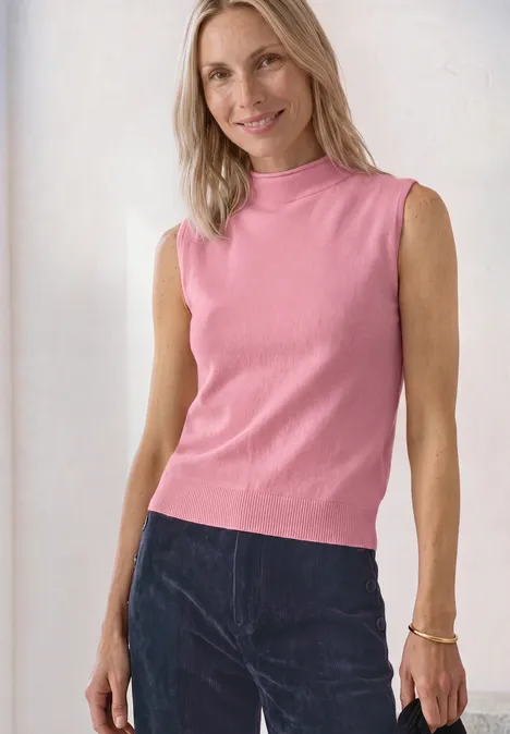 Une femme souriante porte un top rose sans manches et un pantalon foncé.