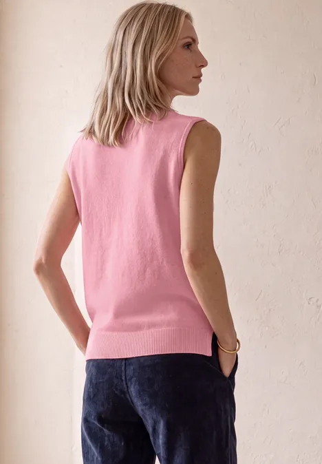 Femme élancée de profil, porte un débardeur rose et un jean foncé, posant devant un fond clair.
