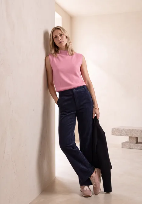 Femme blonde adossée à un mur beige, top rose, pantalon en velours bleu foncé, blazer noir à la main.