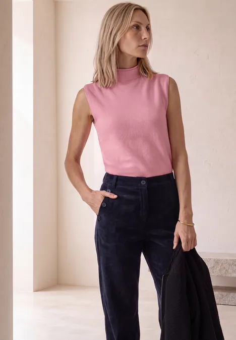 Femme en top rose et pantalon noir, tient une veste noire, regarde de côté.
