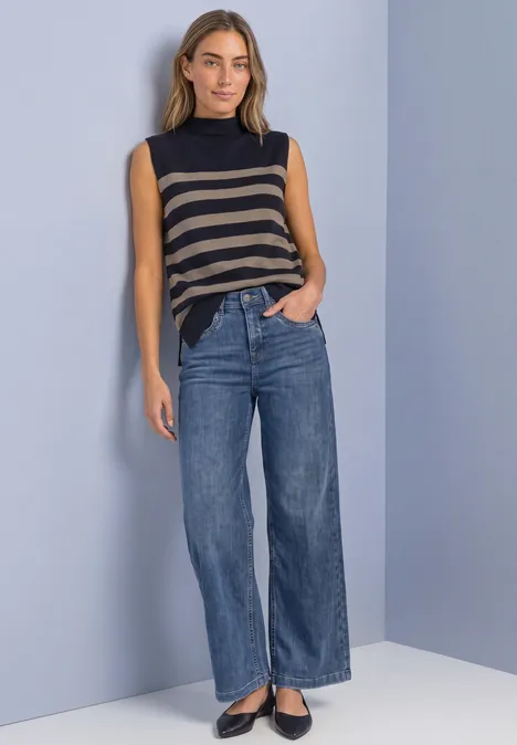 Frau im ärmellosen gestreiften Top, weiten Jeans und schwarzen Ballerinas vor schlichtem Hintergrund.