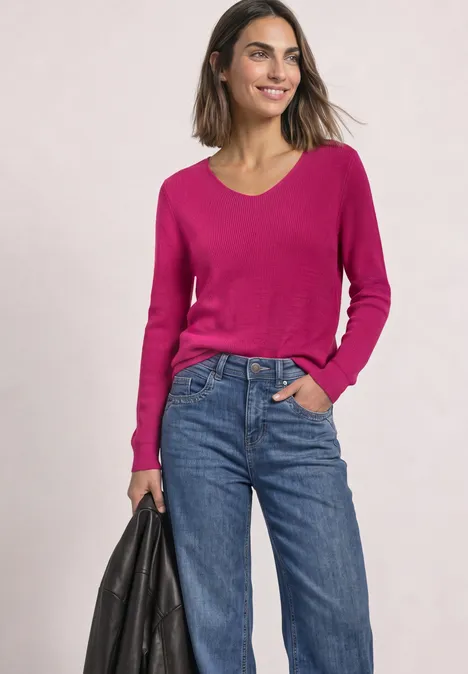 Femme vêtue d’un pull rose et d’un jean bleu, tenant un sac noir et souriant.