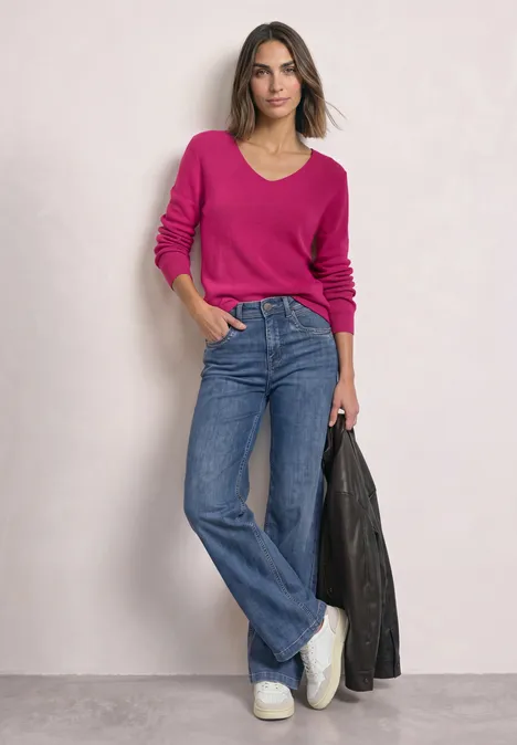 Femme avec pull rose, jean large, baskets blanches, tenant une veste en cuir noire devant un mur clair.