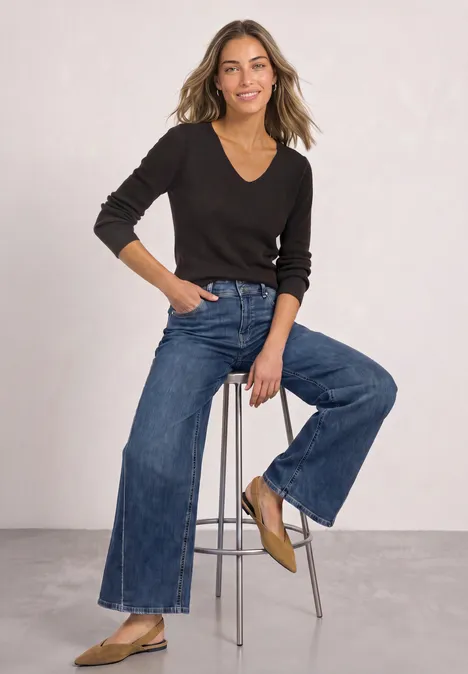 Eine Frau sitzt lächelnd auf einem Hocker, trägt schwarzen V-Pullover, weite Jeans und beige flache Schuhe.