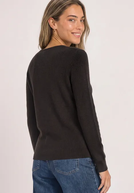Frau in schwarzem Pullover und Jeans blickt über die Schulter, lächelnd vor weißem Hintergrund.