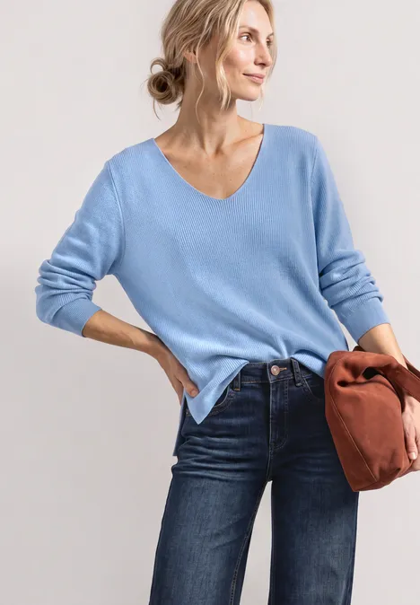 Frau trägt hellblauen V-Pullover und Jeans, hält braune Tasche, lässige Pose.