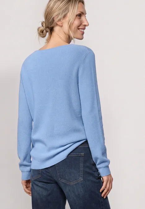 Frau steht mit dem Rücken zur Kamera, trägt hellblauen Pullover und Jeans, lächelt seitlich.