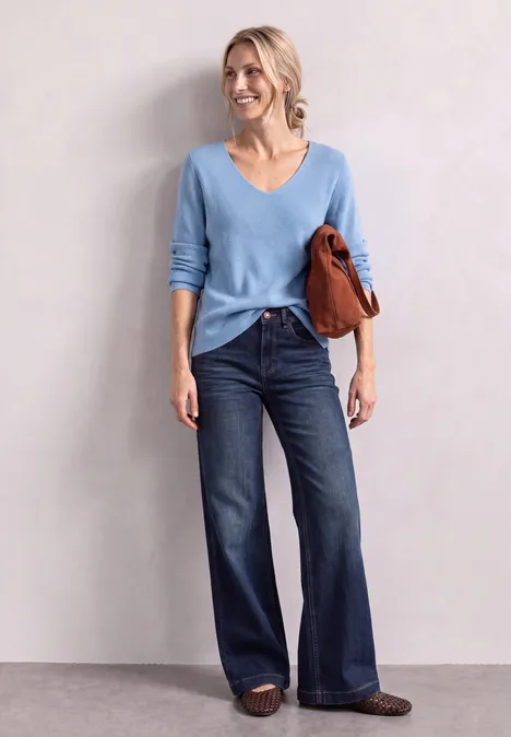 Frau in hellblauem V-Ausschnitt-Pullover und weiten Jeans, trägt braune Sandalen und eine rostbraune Tasche.
