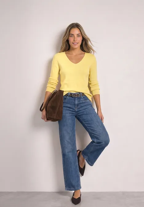 Femme en pull jaune, jean bleu et sac marron souriant devant un fond neutre.