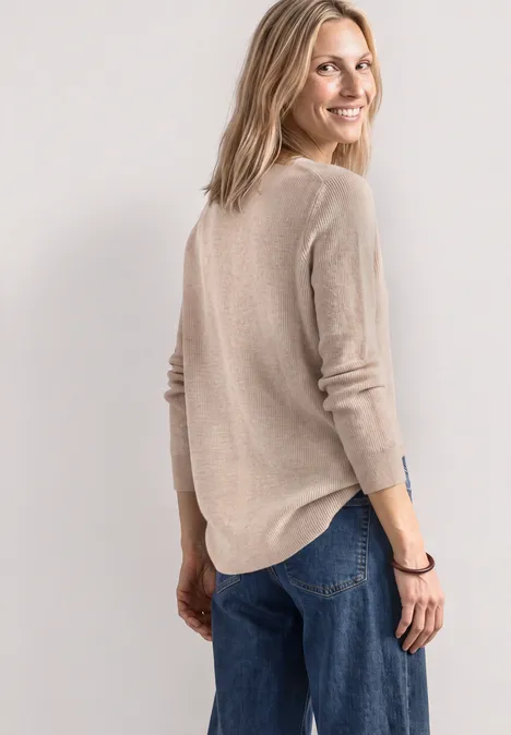 Une femme souriante en pull beige et jean regarde par-dessus son épaule.