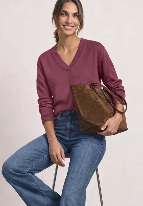 Vrouw zit op kruk, draagt bordeauxroze trui, blauwe jeans en houdt een bruine suède tas vast.