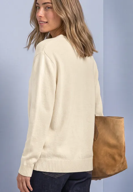 Femme en pull beige portant un sac en cuir marron, regard par-dessus l’épaule sur fond neutre.