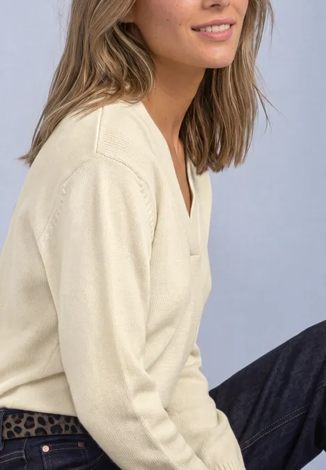 Femme assise de profil, portant un pull col V crème et un jean foncé devant un fond bleu.