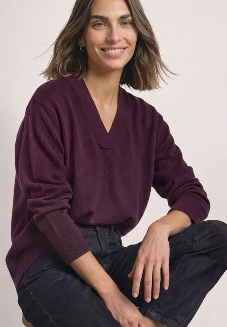 Une femme souriante porte un pull violet à col en V et un jean noir, assise confortablement.