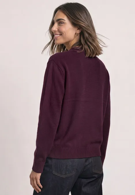 Femme aux cheveux mi-longs portant un pull violet et un jean sombre, souriante en regardant par-dessus son épaule.