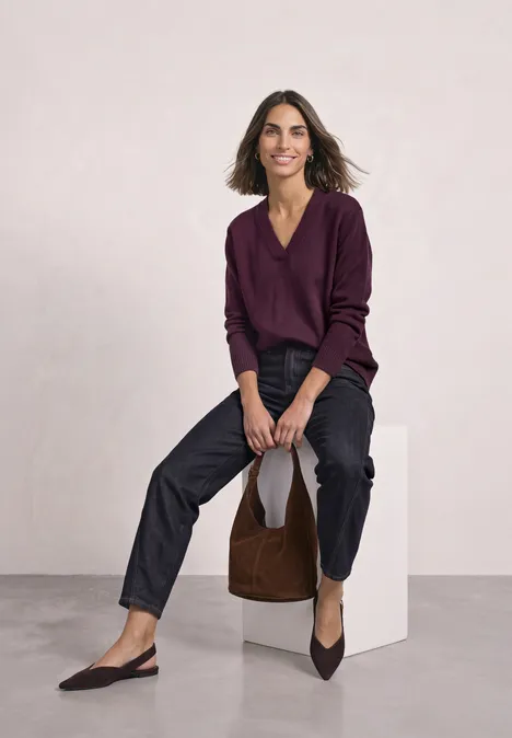 Femme souriante assise sur un cube blanc, pull violet, pantalon noir, ballerines pointues et sac marron.