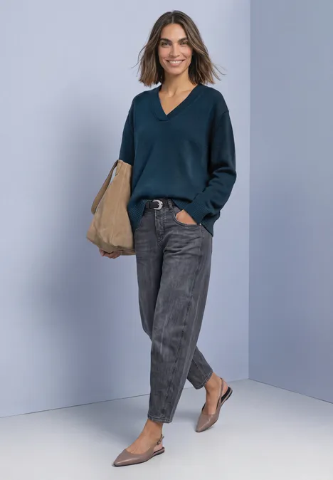 Femme en pull col V bleu foncé, pantalon gris, ballerines beiges et sac, posant devant un mur clair.