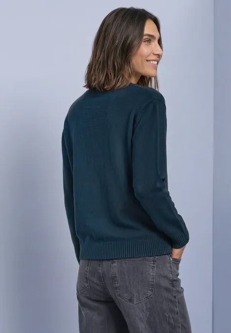 Femme de dos portant un pull bleu foncé et un jean gris, sur fond clair.