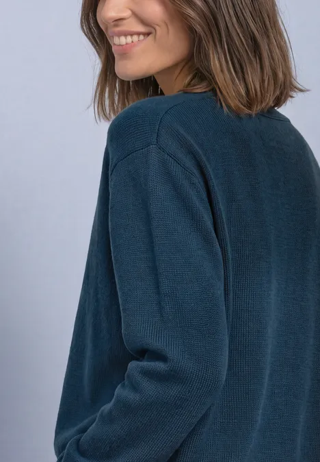 Une femme souriante aux cheveux bruns mi-longs porte un pull vert foncé devant un fond clair.
